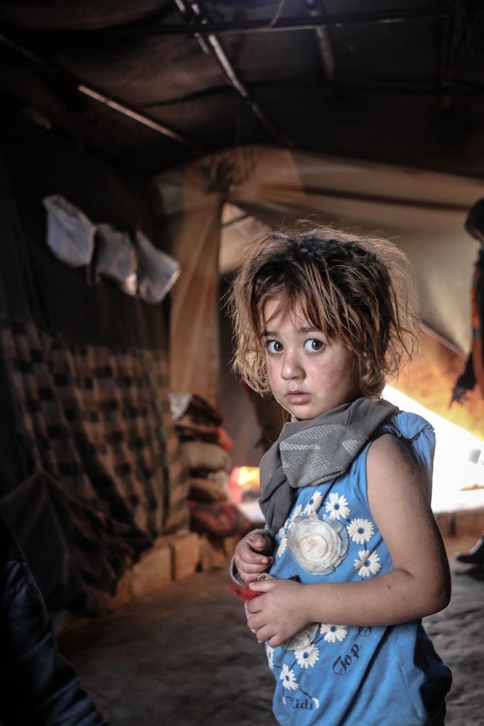 A young girl in a tattered dress looks on with fear inside a dimly lit shelter in Idlib, Syria.